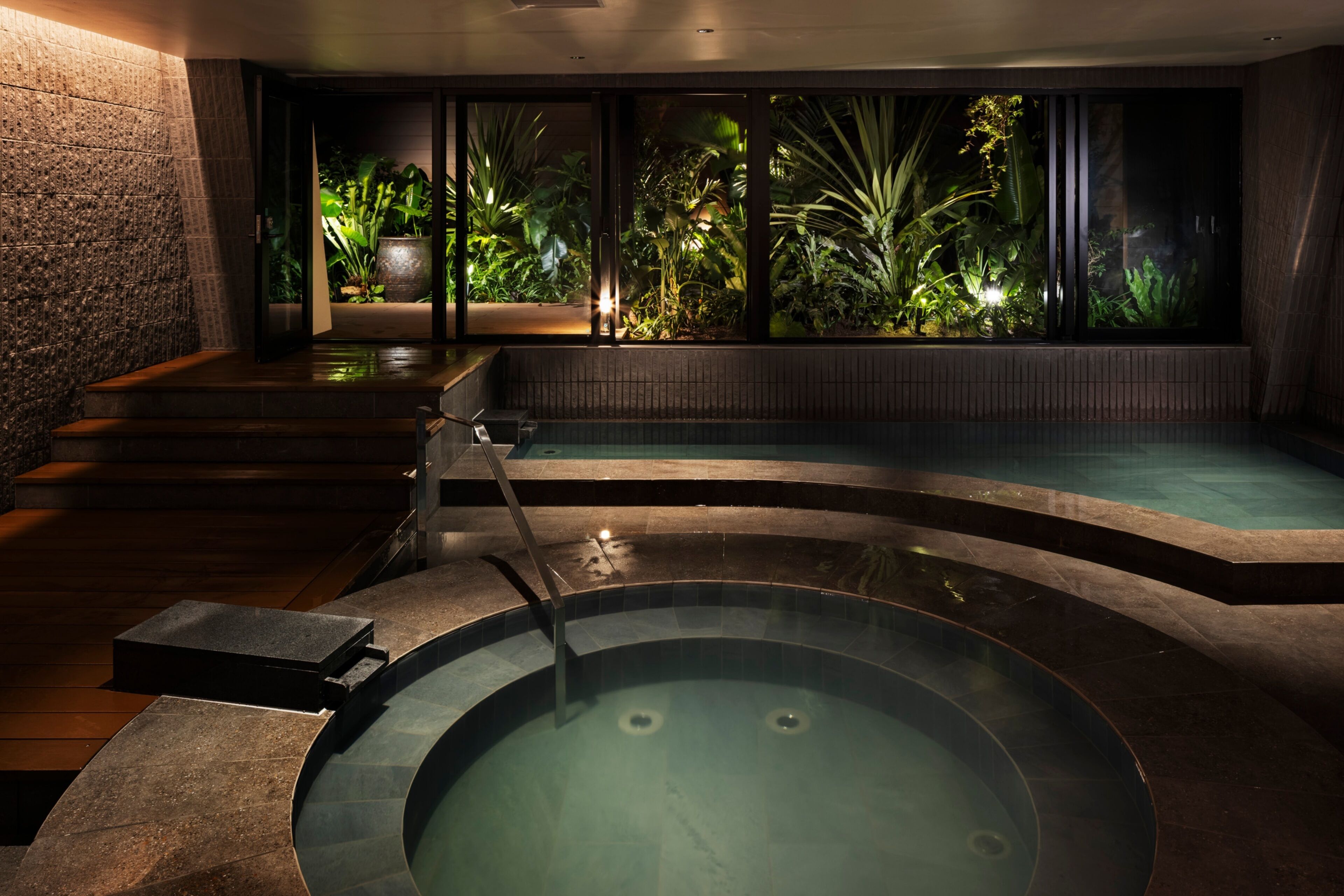 A spa area with a hot tub, a wooden deck, and a view of tropical plants through large windows.