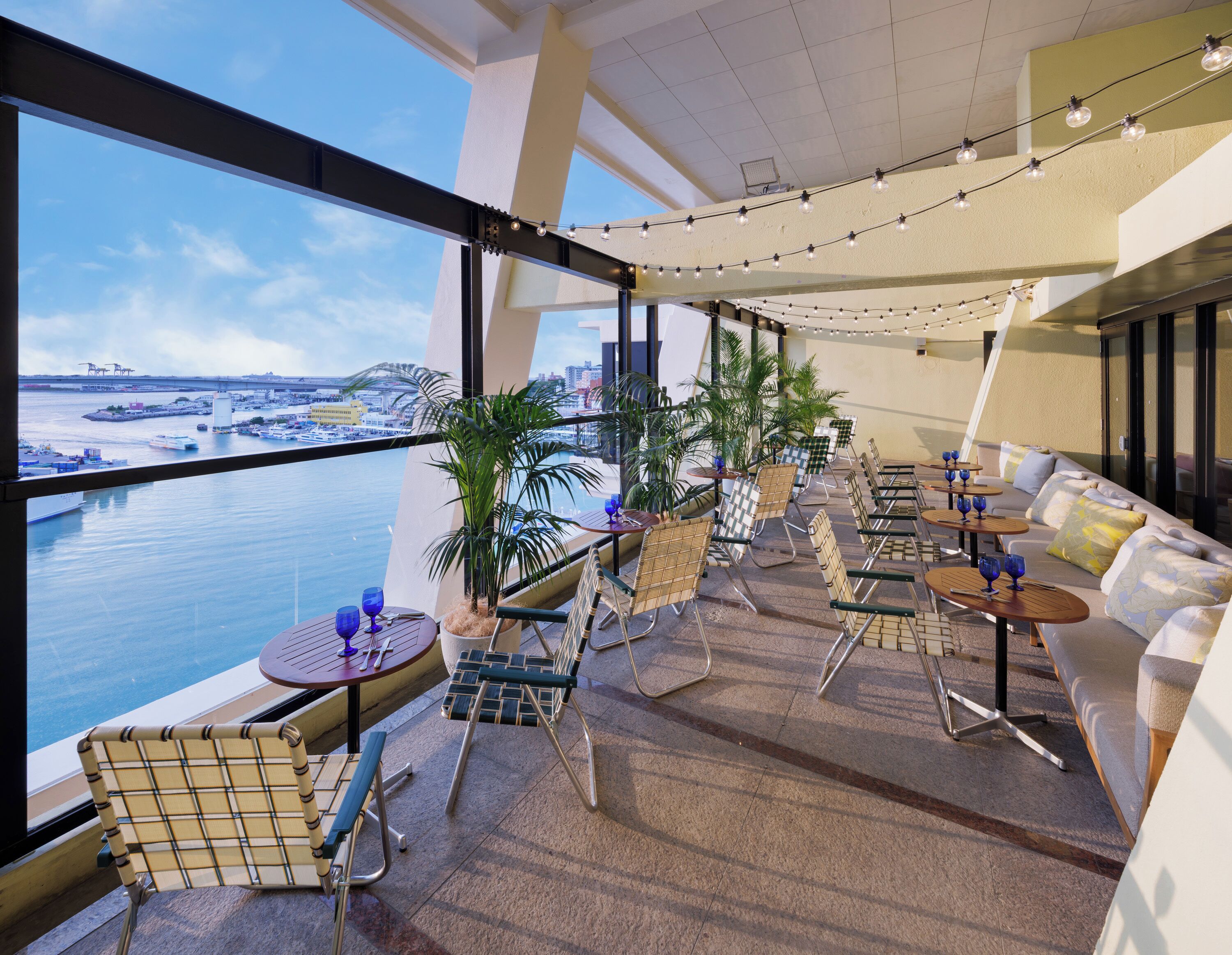 A rooftop terrace with a view of the water, furnished with tables, chairs, and potted plants.
