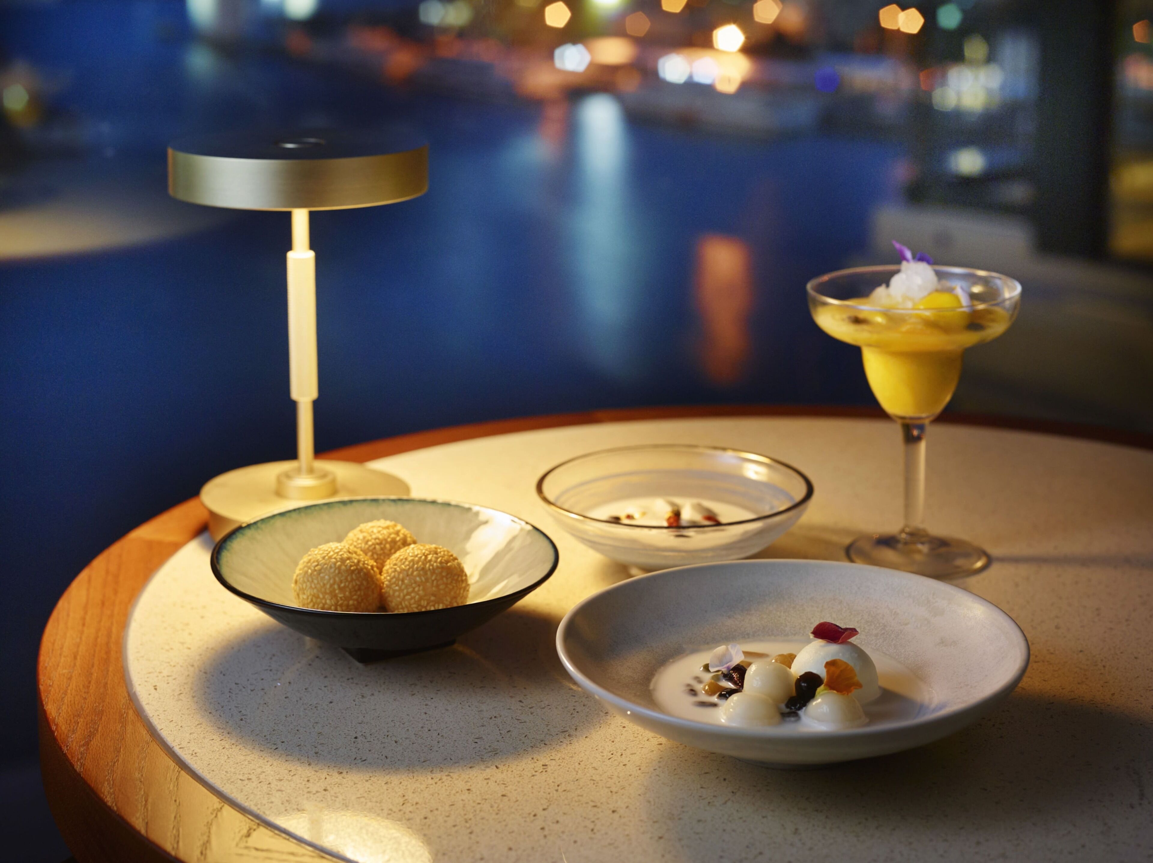 Une table avec trois plats, une lampe et une vue sur une étendue d’eau, de nuit.