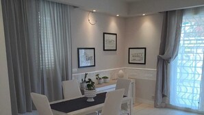 A dining area with a white table and chairs, framed pictures on the wall, and a window with curtains. - Casa Rosa di Sicilia Marzamemi (Marzamemi)