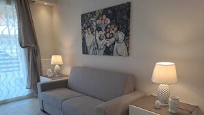 A modern living room with a grey sofa, two bedside tables with lamps, a painting on the wall, and a window with curtains. - Casa Rosa di Sicilia Marzamemi (Marzamemi)
