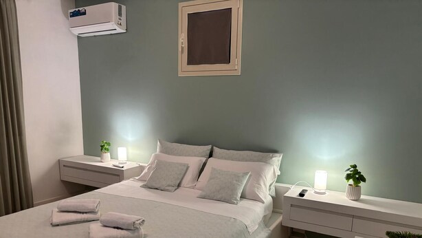 A neatly made bed with pillows and folded towels, a bedside table with a lamp, a small potted plant, and a wall-mounted air conditioner. - Casa Rosa di Sicilia Marzamemi (Marzamemi)