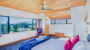 A bedroom with a large window offering a mountain view, a bed with blue and white bedding, a chair, and a wooden bench. - Enjoy 180° views of Lake Tekapo (Lake Tekapo)