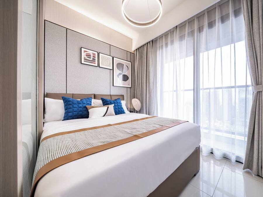 A modern hotel room with a large bed, a headboard with built-in speakers, and a large window with sheer curtains.