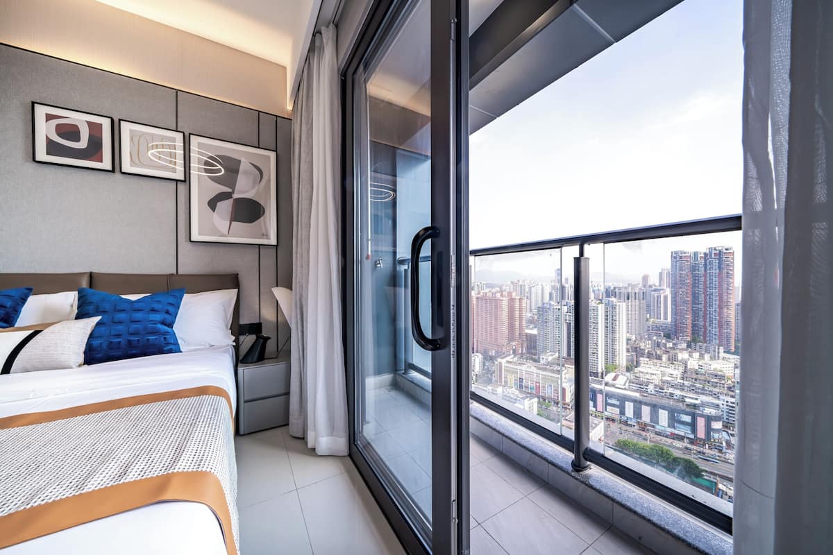 A modern hotel room with a large window offering a city view, a neatly made bed with blue and white bedding, and abstract art on the wall.
