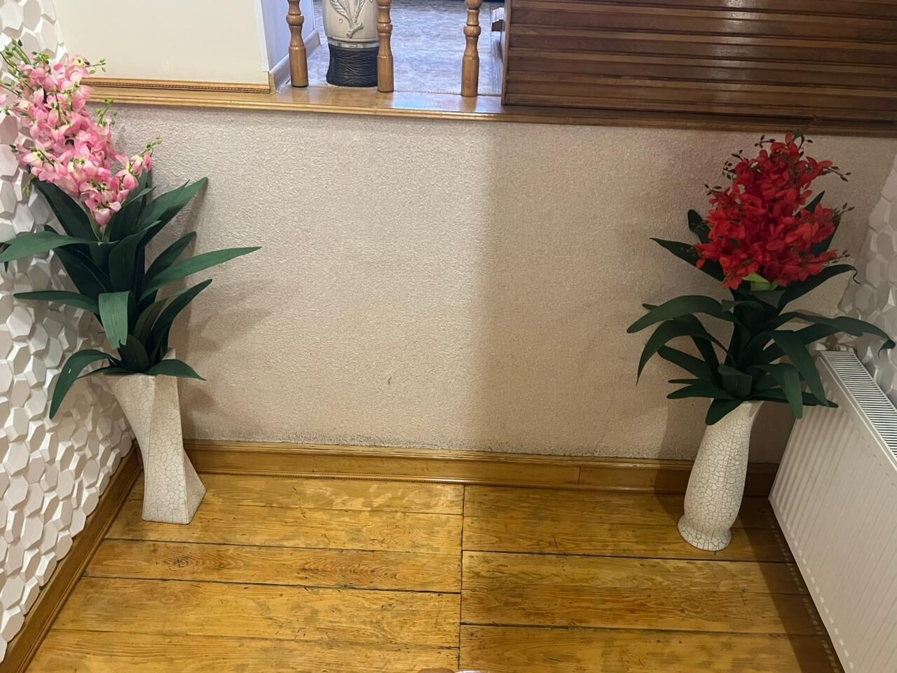 Een hoek van een kamer met houten vloer, twee witte, geribbelde vazen met planten, een houten kast en een witte radiator.