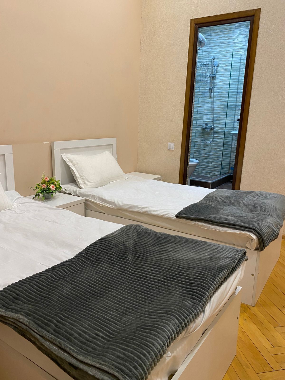 A bedroom with two beds, a small bedside table with a plant, and a glass shower door.