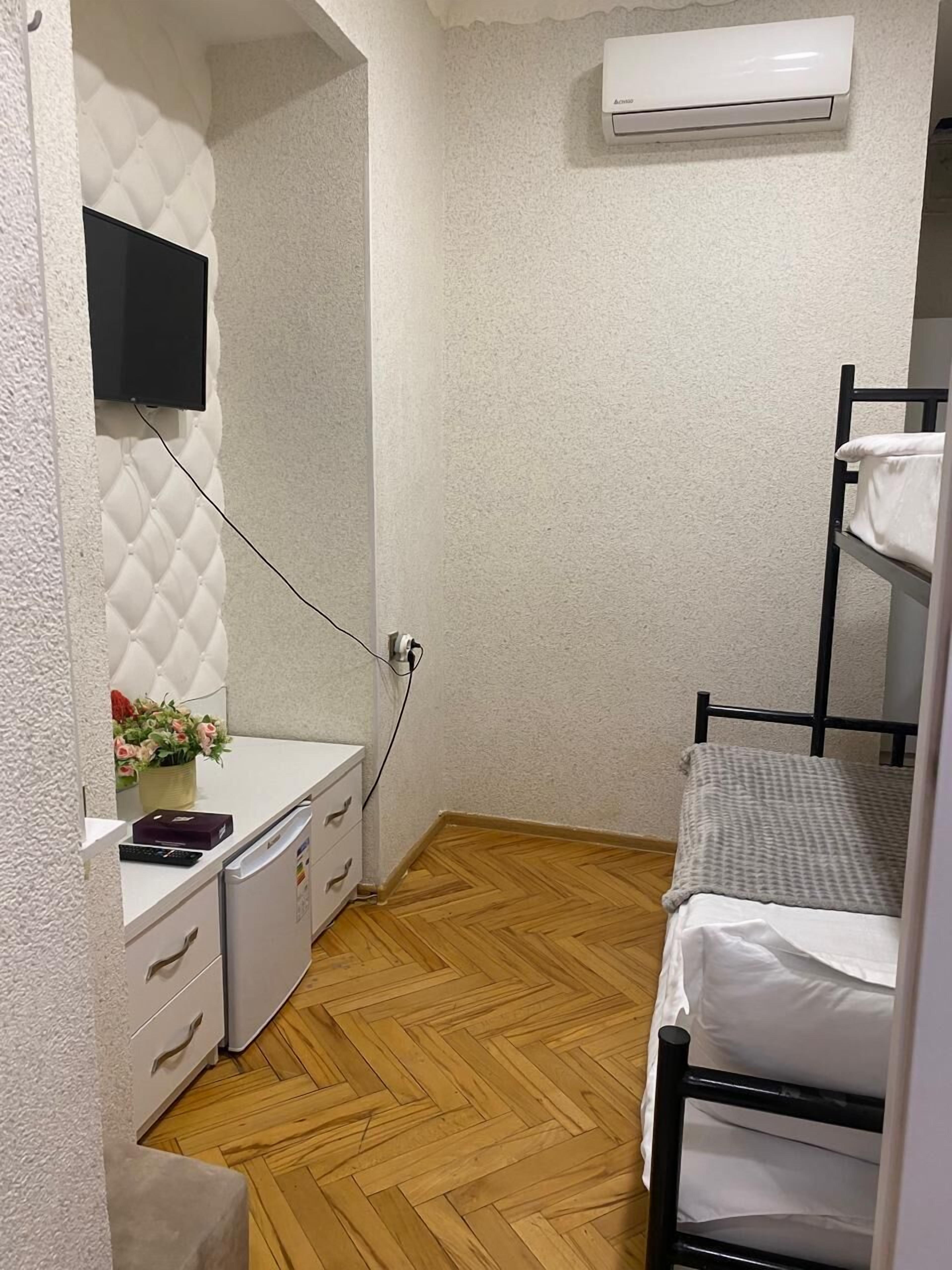 A small, compact room with a bunk bed, a white dresser with drawers, a small white table with a potted plant, and a mounted TV.