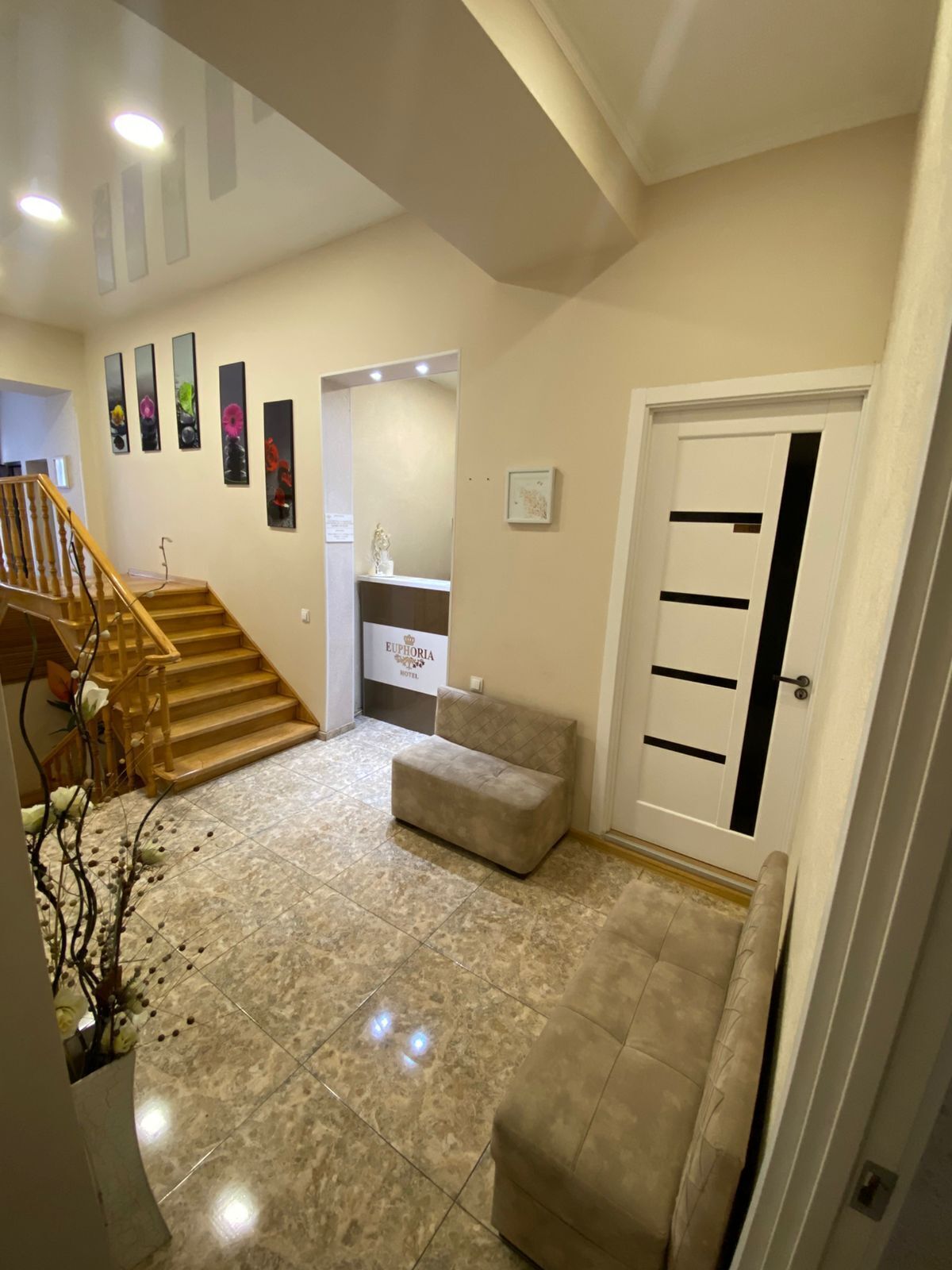 A hotel lobby with a wooden staircase, a cushioned seating area, a reception desk, and a door with a black stripe.