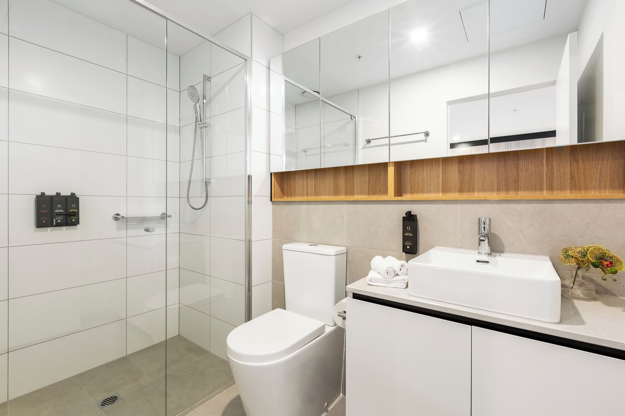 Een moderne badkamer met een douchecabine van glas, een toilet, een wastafel met spiegel erboven en een houten plank.