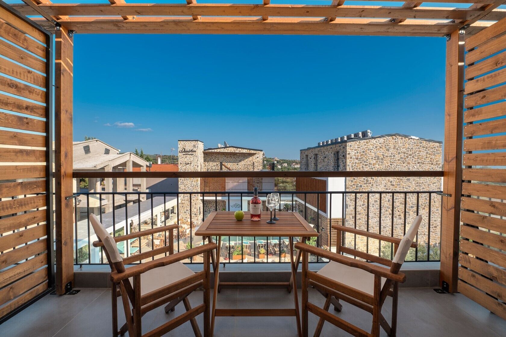 Ein Balkon mit Holztisch und -stühlen, einer Weinflasche und einem Glas. Der Balkon bietet Blick auf ein Steinhaus und einen Pool.