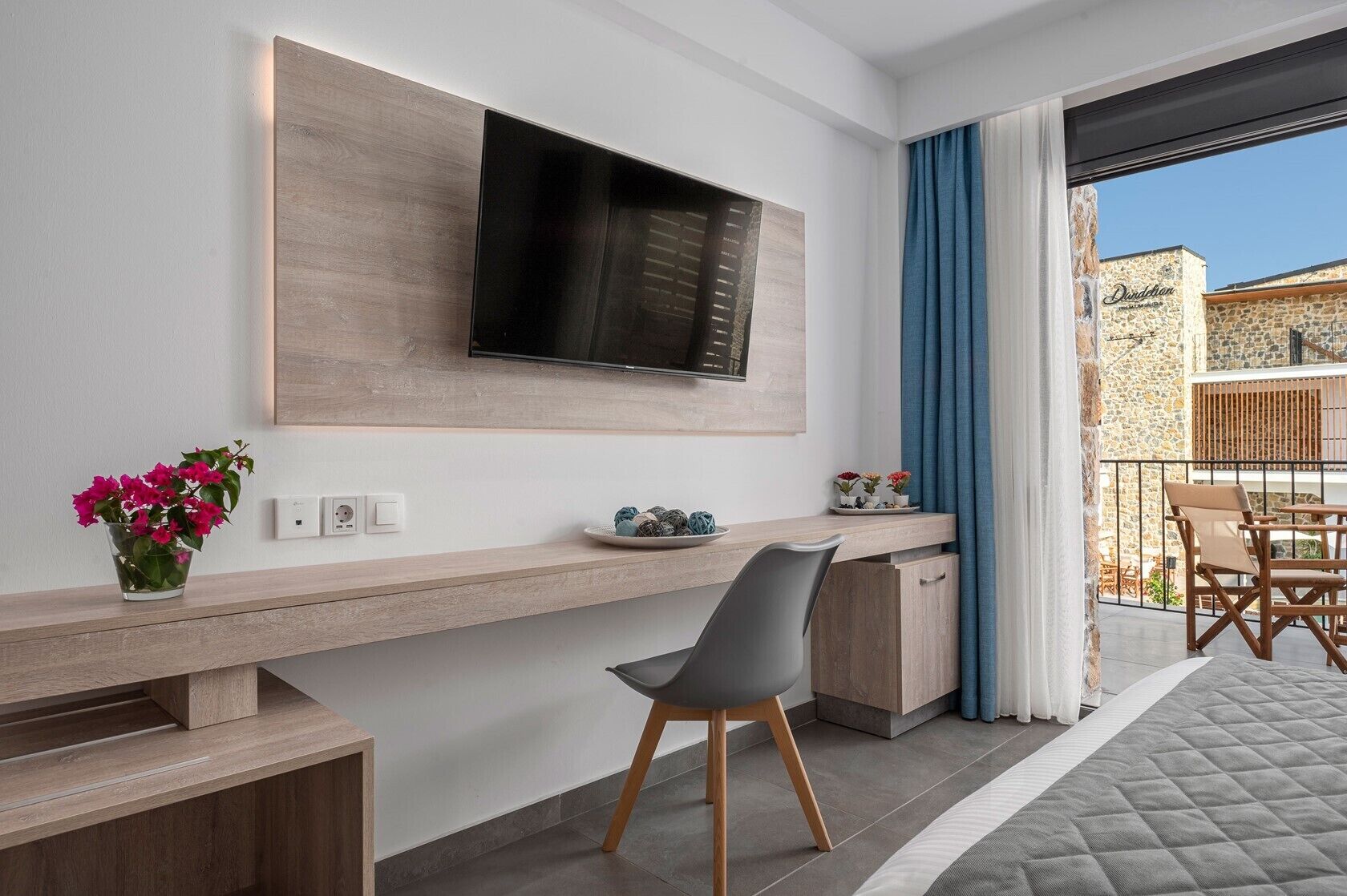 A modern room with a flat-screen TV mounted on a wooden wall, a desk with a chair, and a balcony with a view of a building.