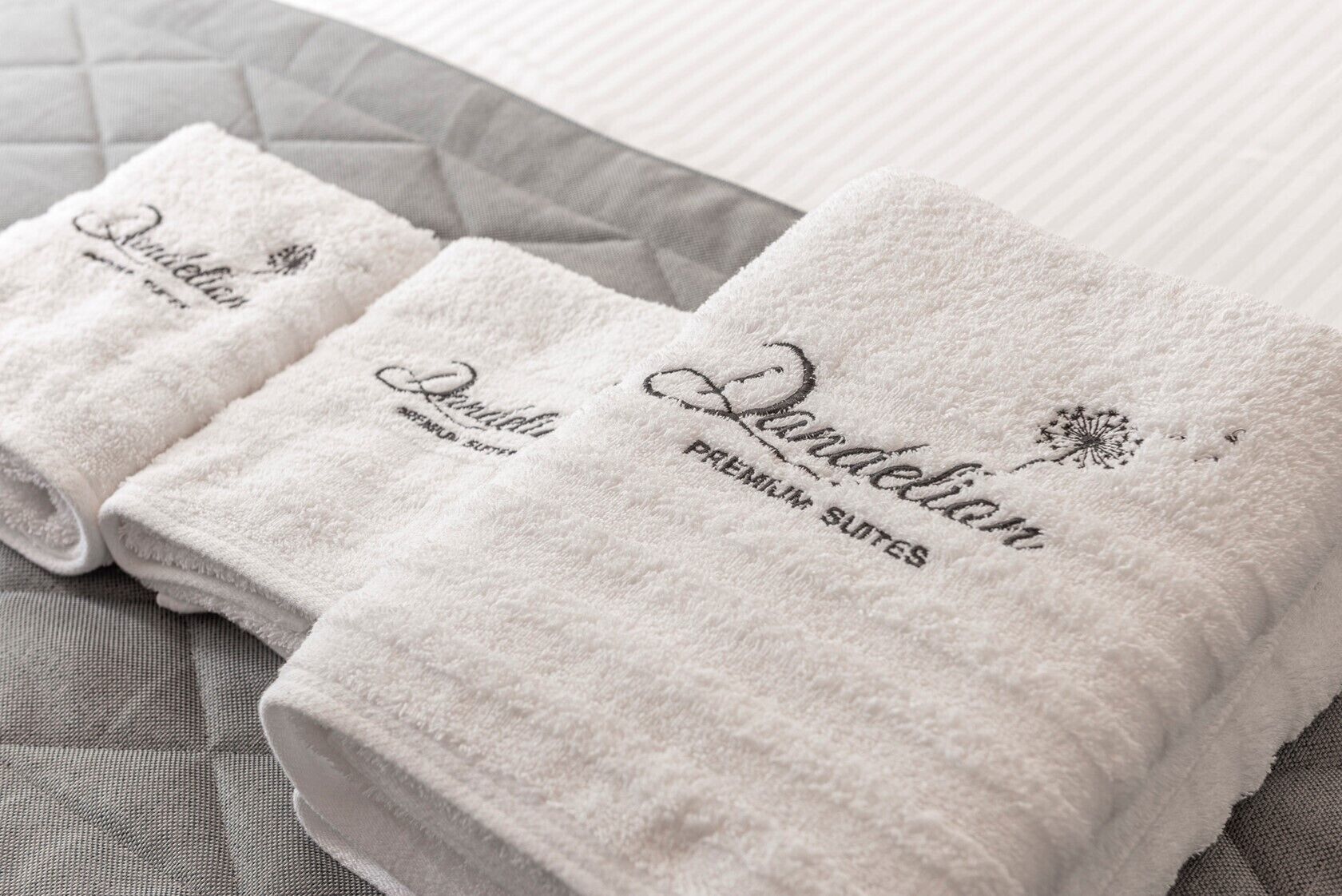 Three neatly folded white towels with "Dandelion Premium Suites" embroidered on them, placed on a textured grey bedspread.