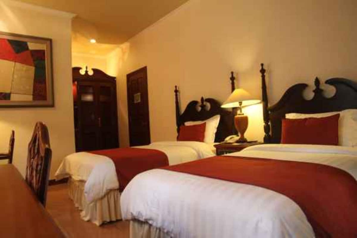 A hotel room with two single beds, a wooden headboard, a bedside table with a lamp, and a framed picture on the wall.