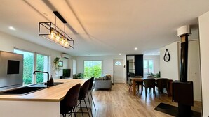 A modern kitchen with a central island, a dining area with a wooden table and chairs, and a large window providing a view of greenery.