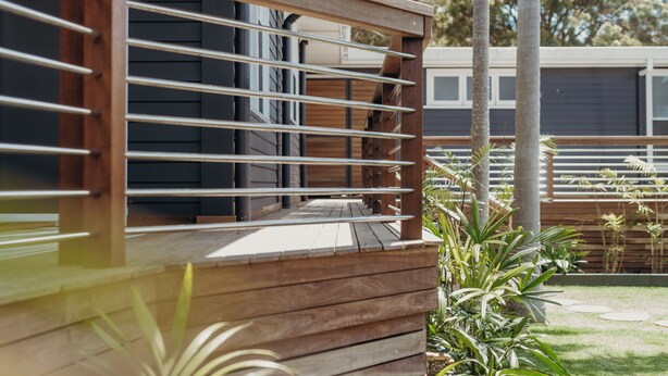 Una terraza de madera con barandas de metal, rodeada de follaje y un edificio moderno al fondo.
