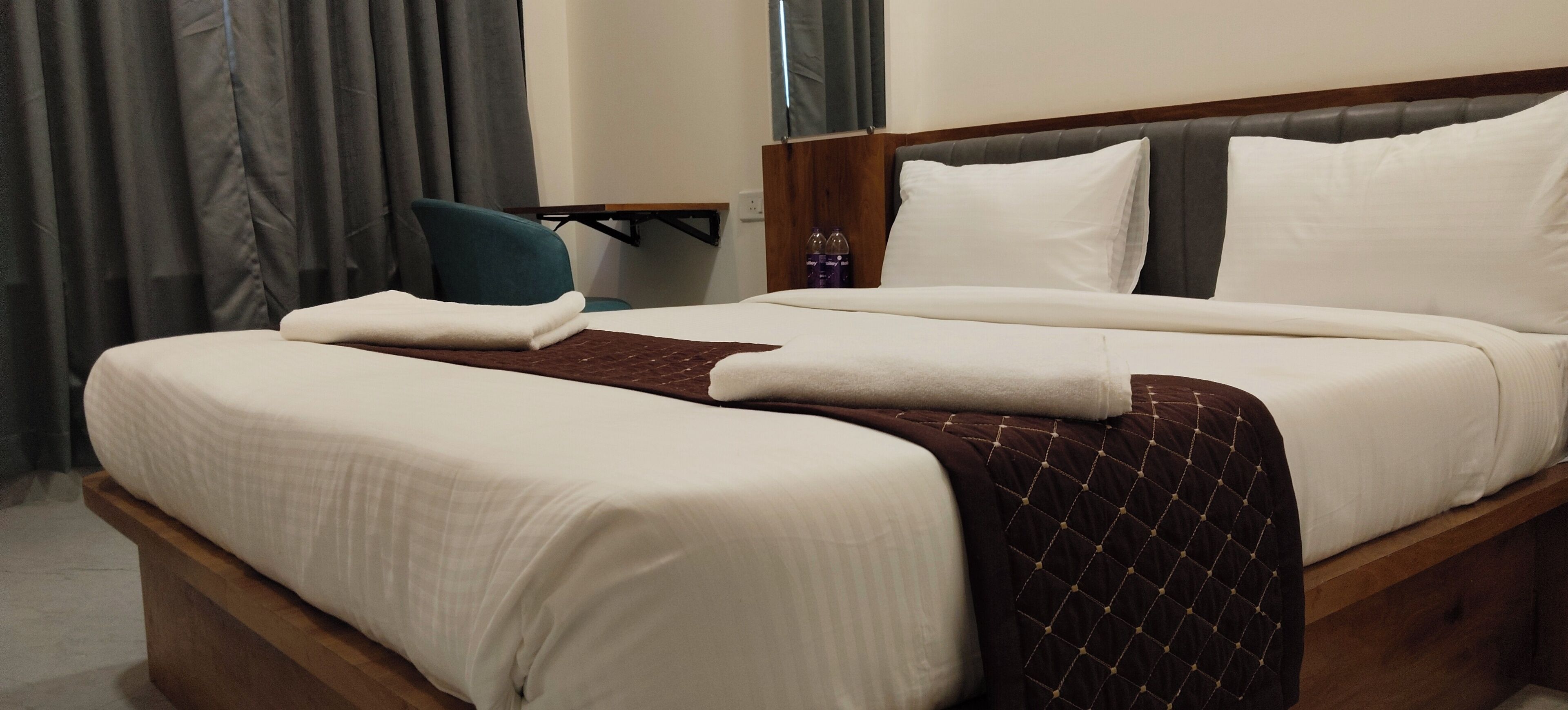 A neatly made bed with a brown quilt, a wooden headboard, and a small table with bottles.