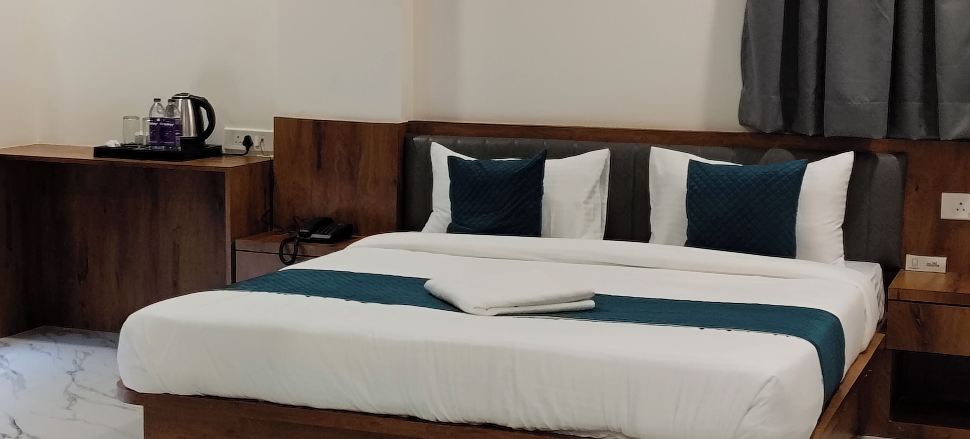 Een net opgemaakt bed met wit en teal beddengoed, een houten hoofdbord en een nachtkastje met een koffiezetapparaat en glazen.