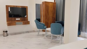 A hotel room with a TV mounted on the wall, two blue chairs, a wooden wardrobe, and a marble floor. - Hotel Sai Executive (Rajgurunagar)