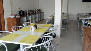 A dining area with white metal chairs and tables, a wooden cabinet with stacked plates, and a shelf with pots and pans. - Hotel Sai Executive (Rajgurunagar)