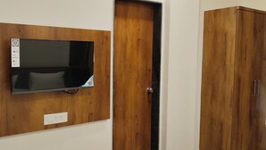 A room with a mounted HD TV on a wooden panel, a door with a black handle, and a wooden wardrobe. - Hotel Sai Executive (Rajgurunagar)