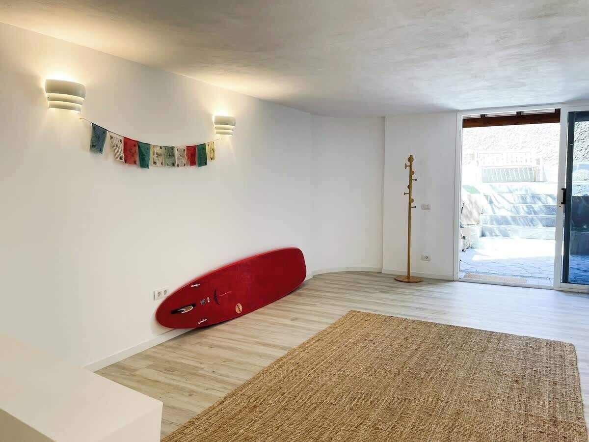 Um quarto com uma prancha de surf vermelha encostada na parede, um abajur e uma janela grande.
