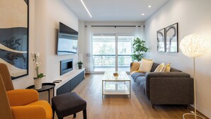 Una sala de estar moderna con un sofá, una mesa de centro, un televisor montado en la pared y un gran ventanal que proporciona luz natural.