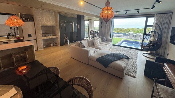 A spacious living area with a sofa, dining table, and chairs, a kitchen with a stone backsplash, and a large window offering a view of the outdoors.
