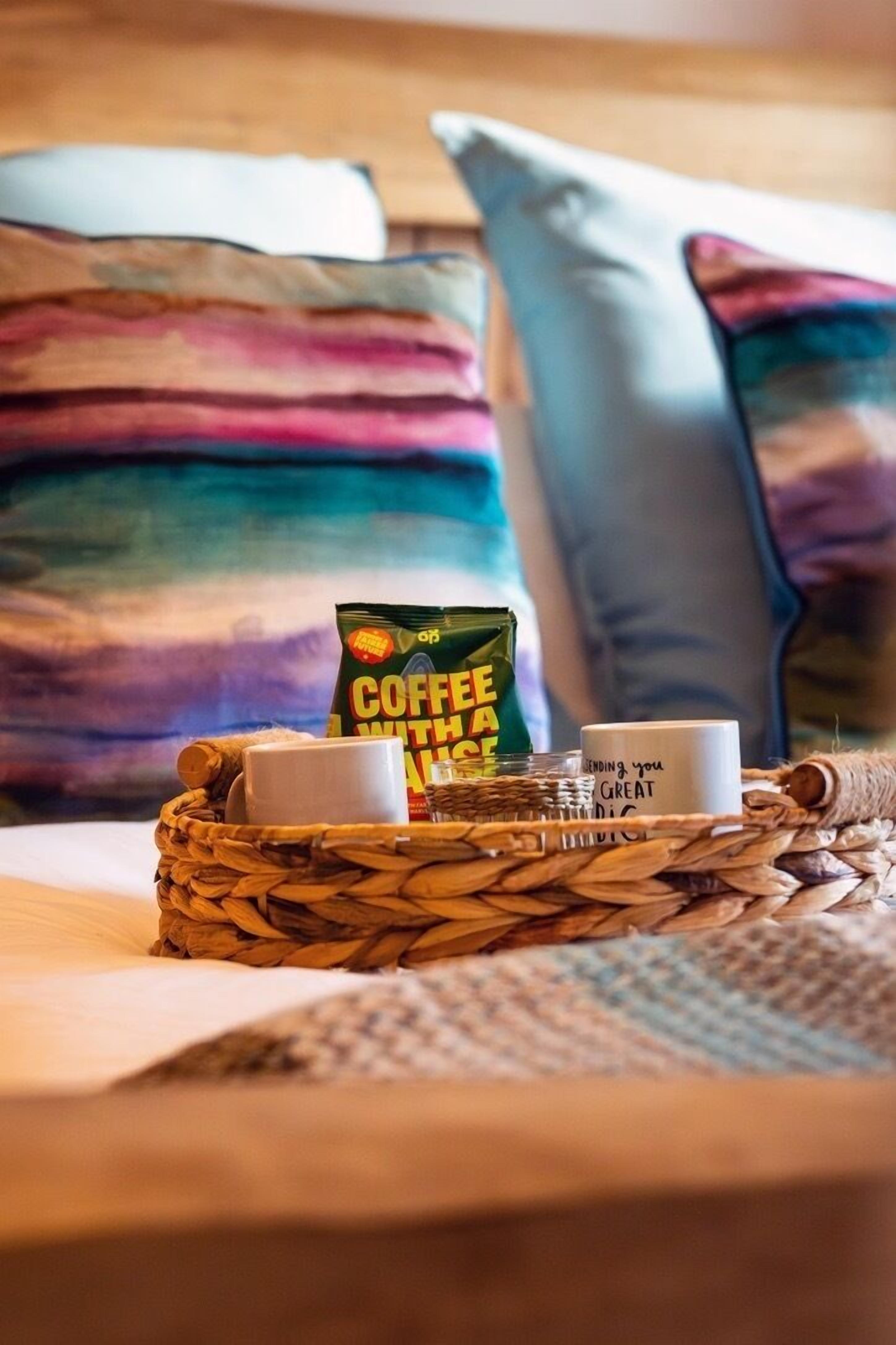 A bed with a woven basket containing a coffee packet, two white mugs, and a candle with a message.