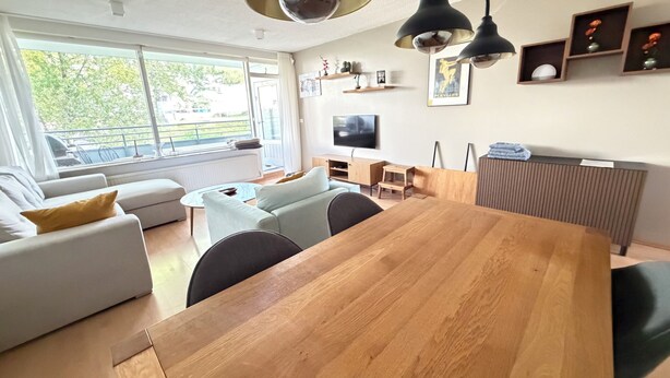 A modern living room with a wooden dining table, white sofas, and a large window with a view of greenery. - Cosy and family friendly apartment in Kópavogur - Birta Rentals (Kópavogur)