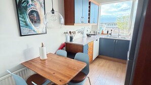 A modern kitchen with wooden cabinets, a dining table with chairs, a hanging light fixture, and a window with a view of the city. - Cosy and family friendly apartment in Kópavogur - Birta Rentals (Kópavogur)