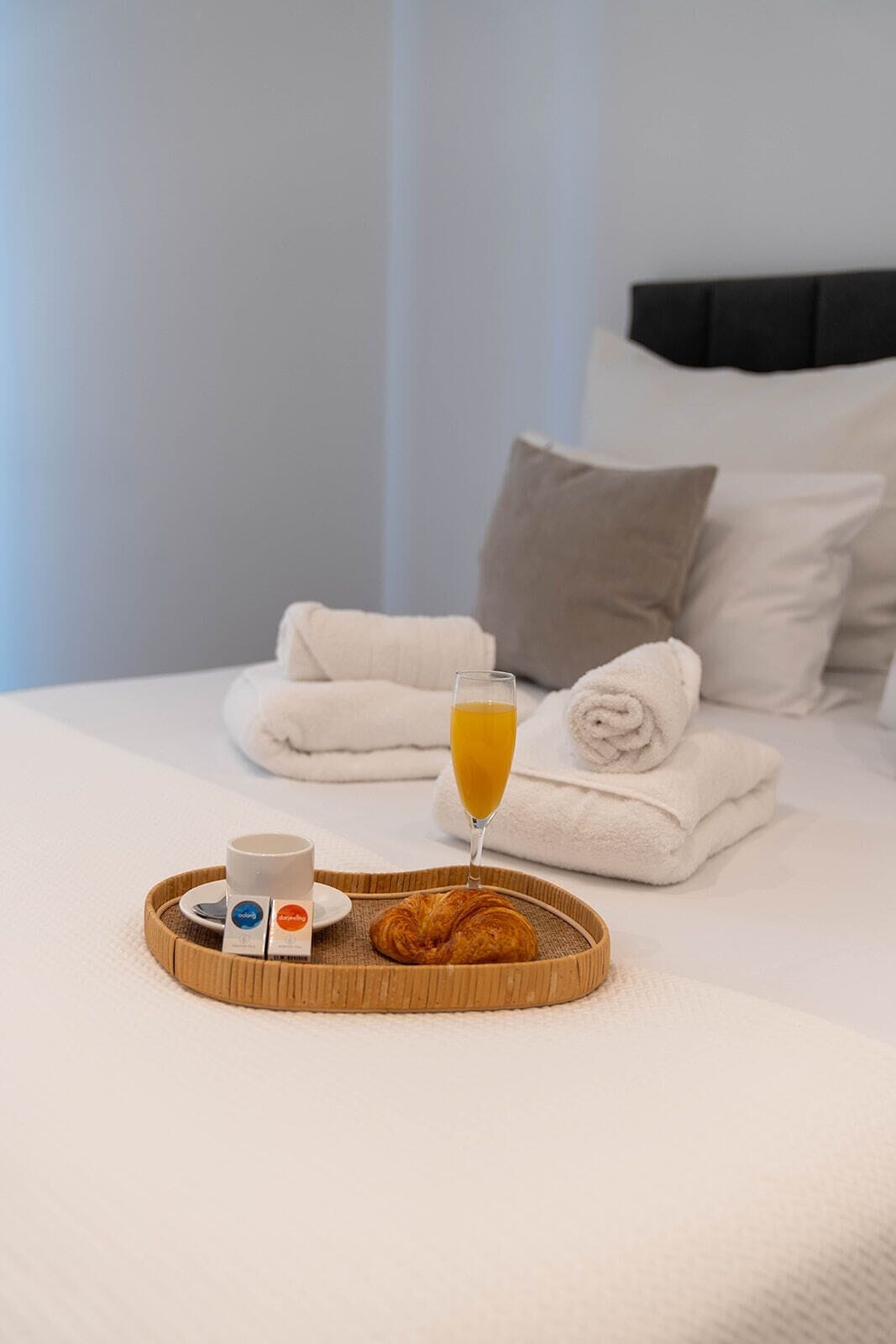 Un lit parfaitement fait, avec du linge de lit blanc et des serviettes pliées, un plateau avec des croissants, un verre de jus d’orange et deux tasses de café.