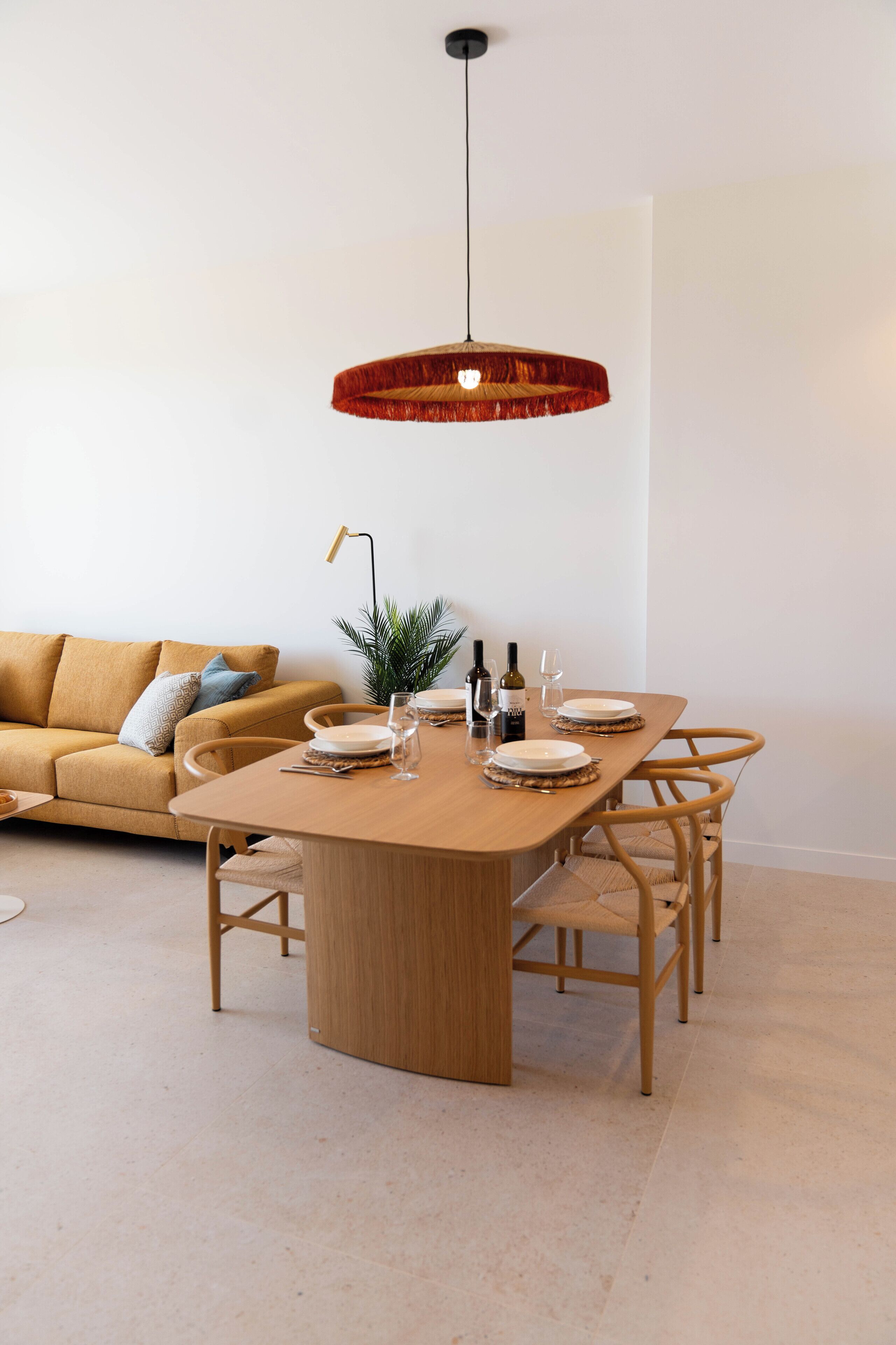 A dining area with a wooden table set for four, surrounded by chairs. A hanging light fixture is above the table.