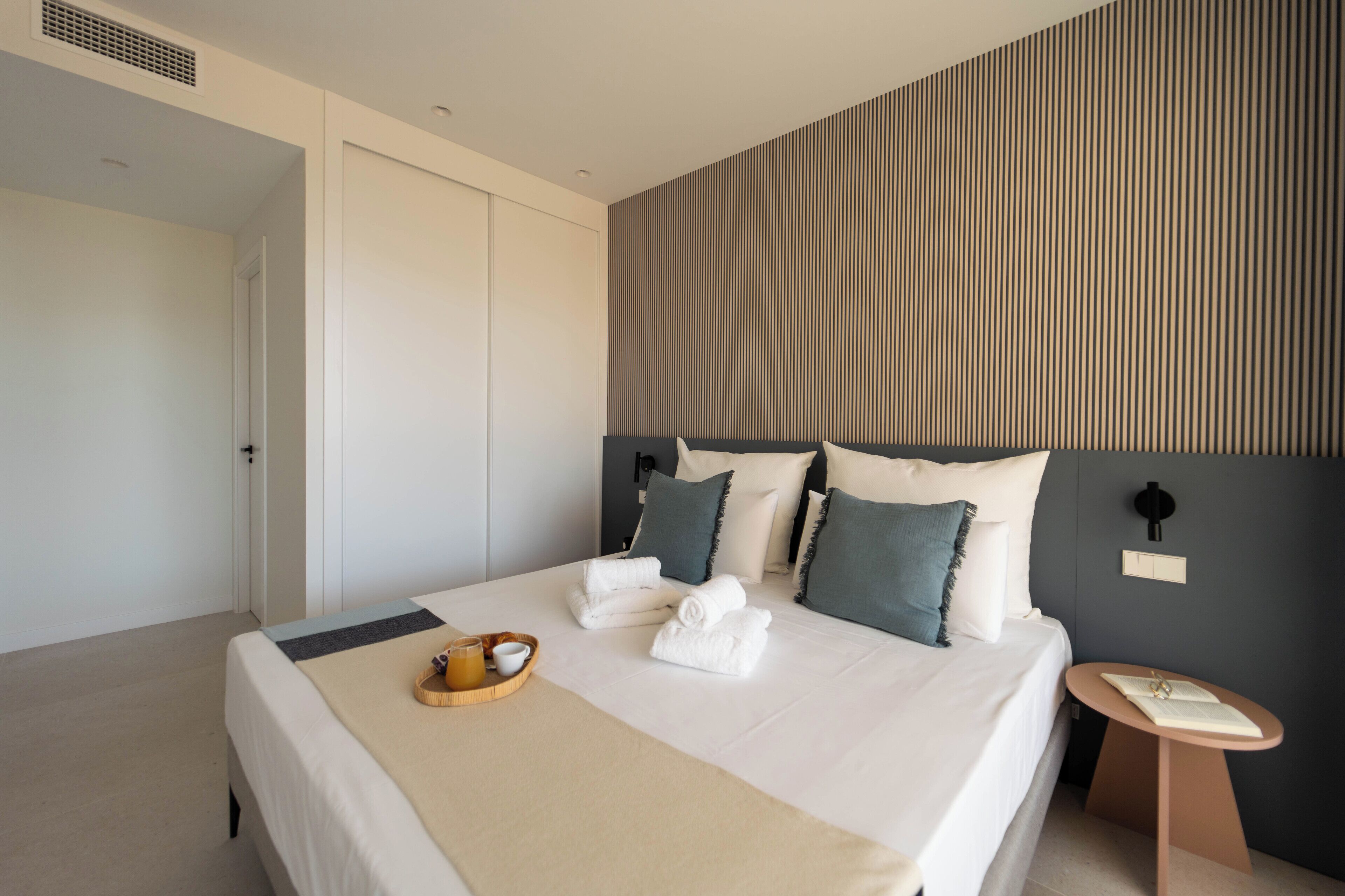 A modern hotel room with a neatly made bed, a bedside table with a lamp, and a wall-mounted light fixture.