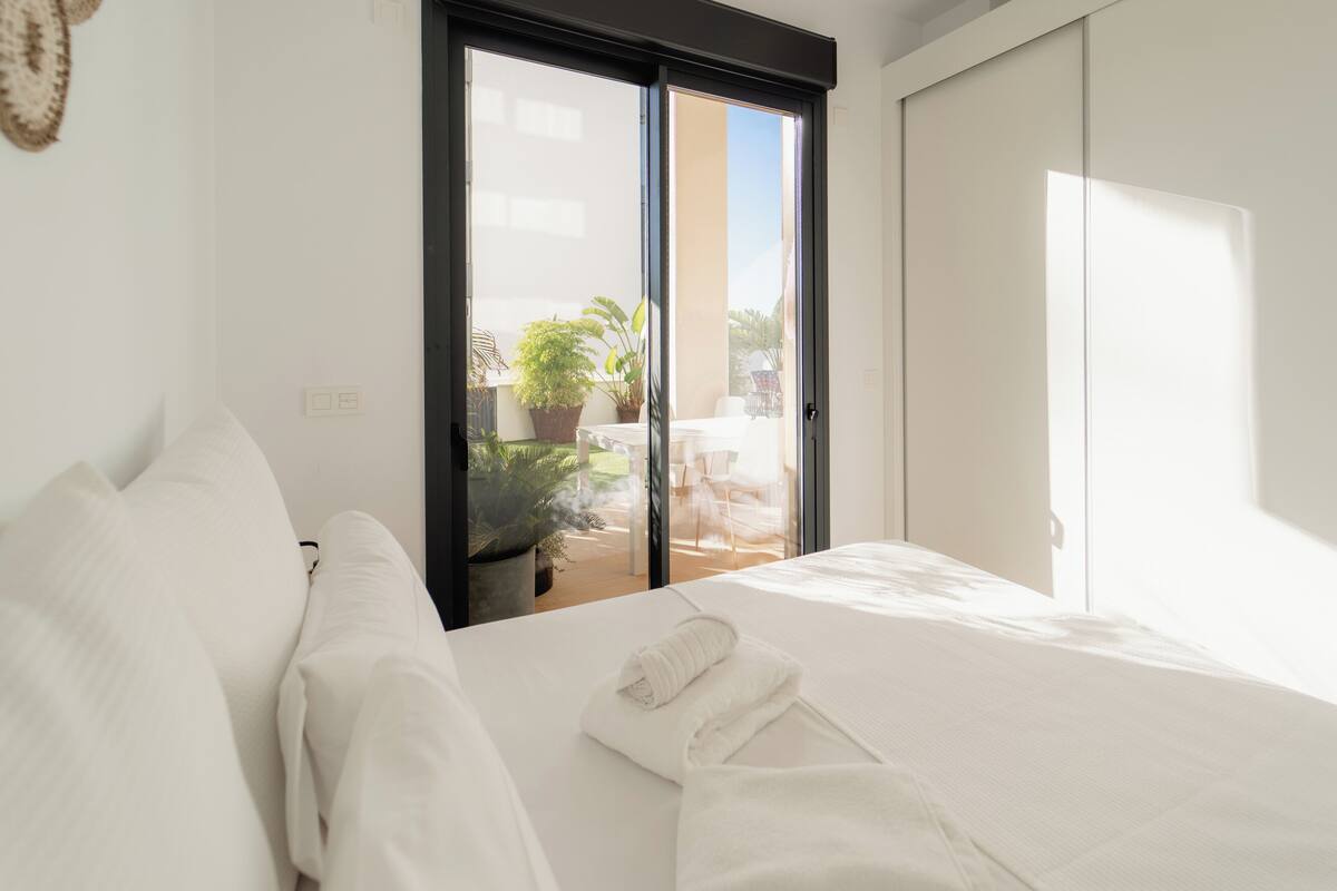 A hotel room with a bed, white linens, a sliding glass door to a balcony with a view of the outdoors, and a potted plant.