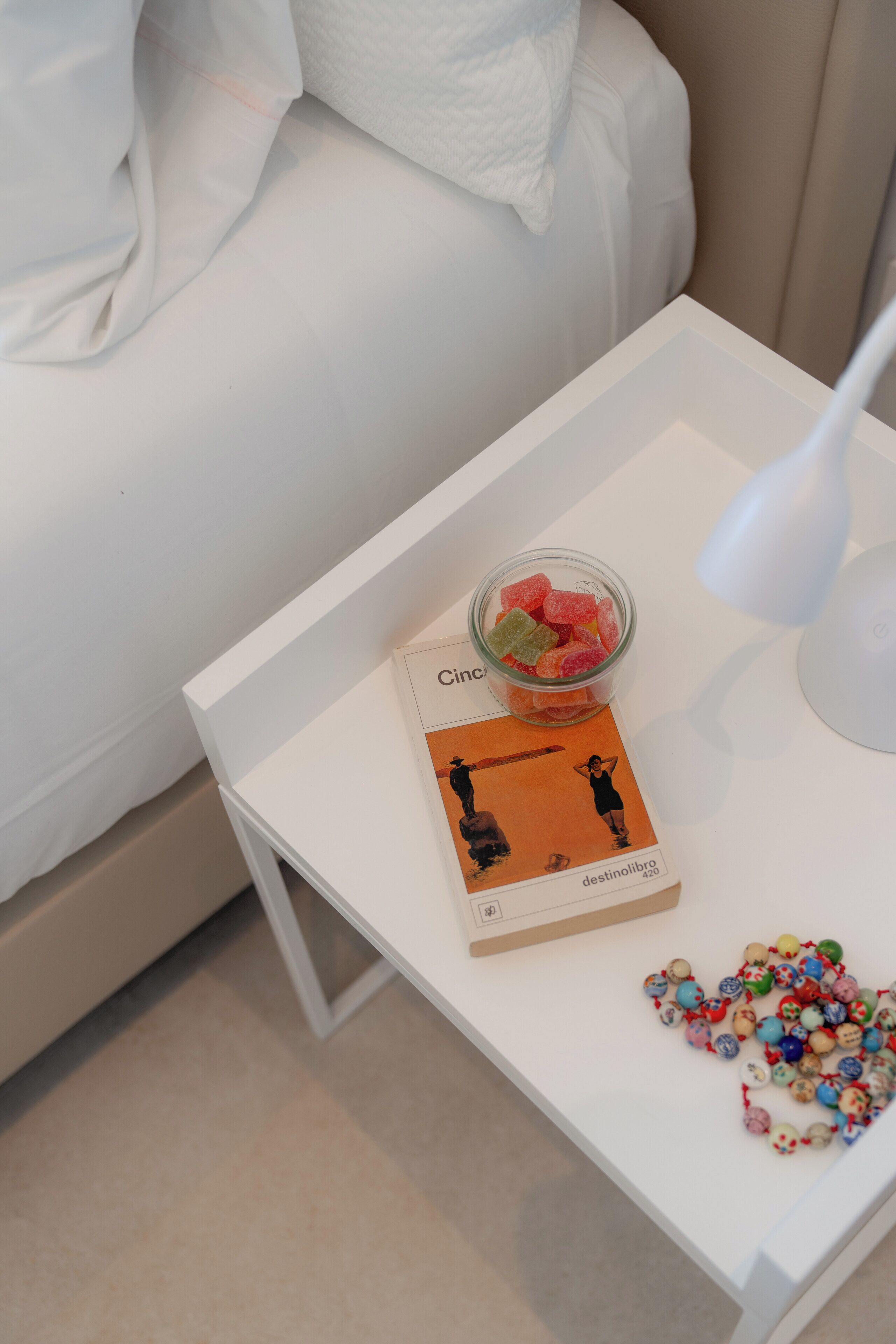 A white bedside table with a book, a jar of colorful candies, and a white lamp.