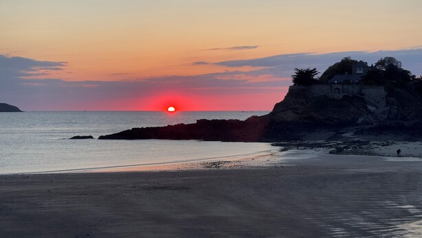 A beach with a sunset, rocky coastline, and a building on a cliff.