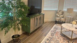 Una sala de estar con un aparador verde, un televisor, un sillón estampado, una pequeña mesa redonda con una lámpara y una planta grande en maceta.