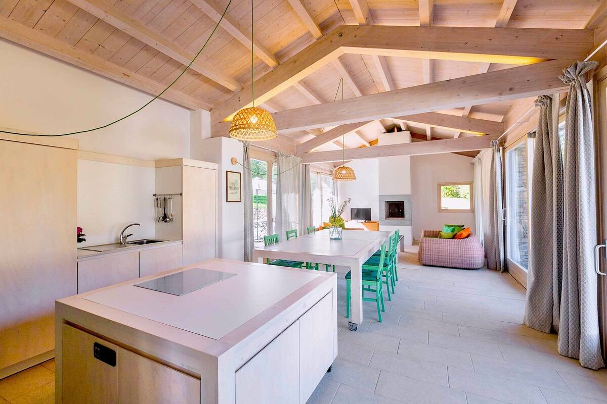 A modern kitchen with a central island, a dining area with green chairs, and a cozy seating area with a cushion.