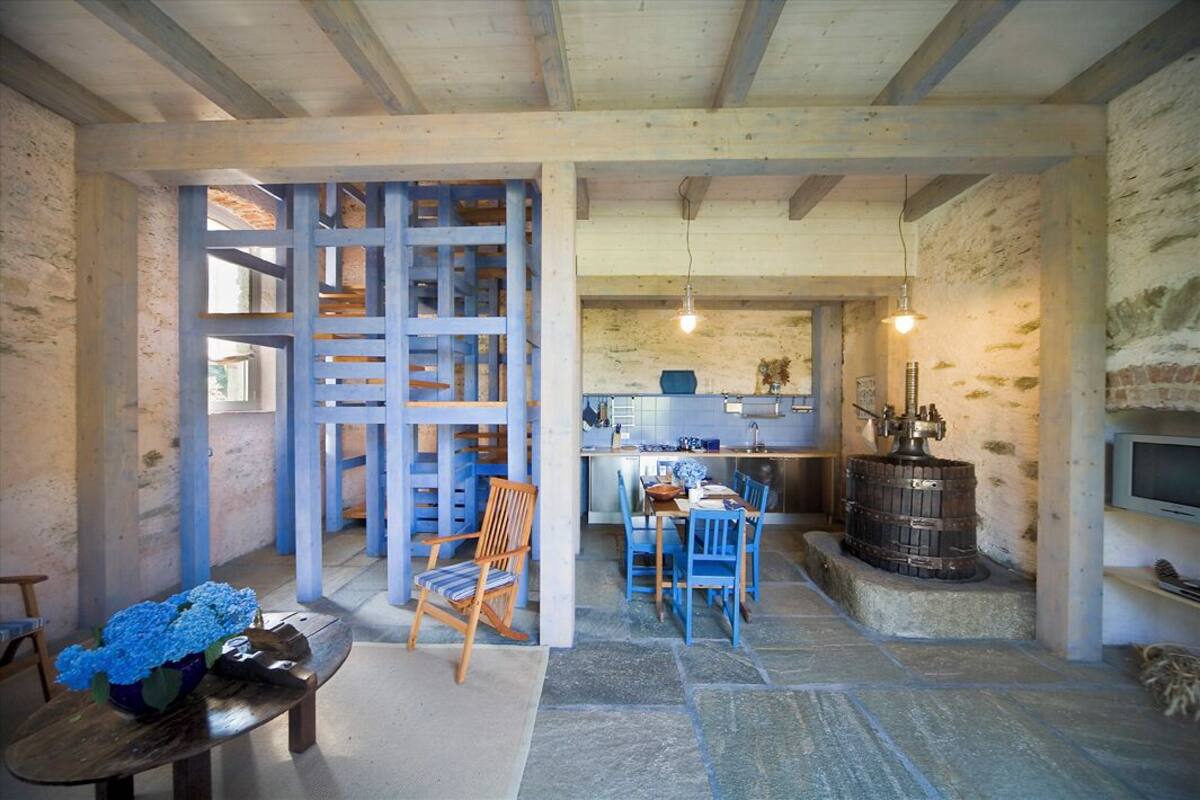 A rustic interior with exposed wooden beams, a stone floor, a dining area with blue chairs, and a kitchen with a large stove.