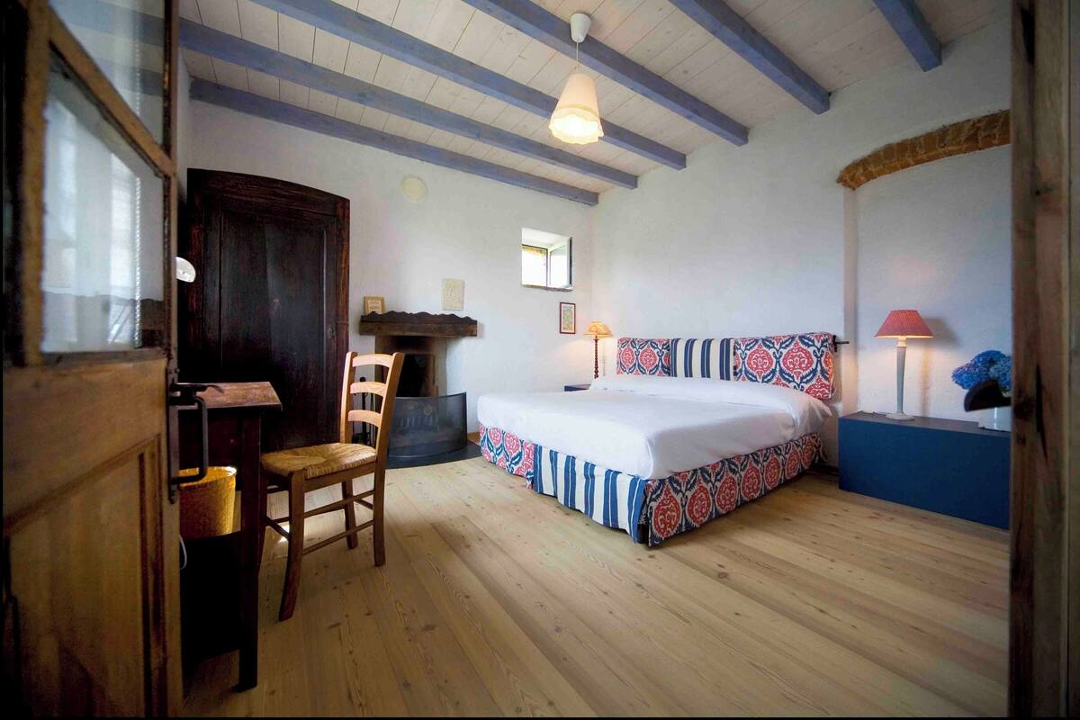 A bedroom with a wooden floor, a bed with patterned bedding, a chair, a desk, a fireplace, and a lamp.