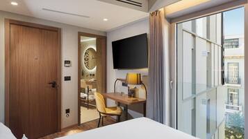 A hotel room with a bed, a desk with a lamp, a TV, a wooden door, and a view of the outside.