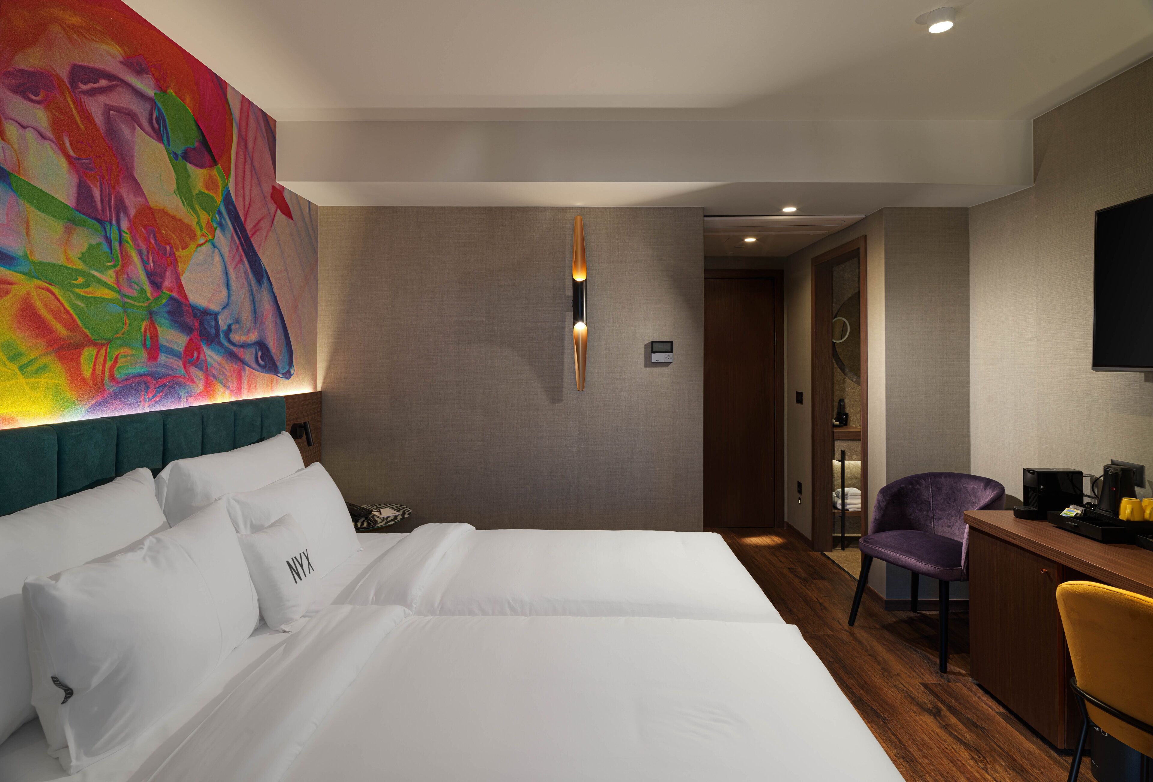 Ein Hotelzimmer mit einem großen Bett, einem Schreibtisch, einem Stuhl und einem farbenfrohen, abstrakten Gemälde an der Wand.