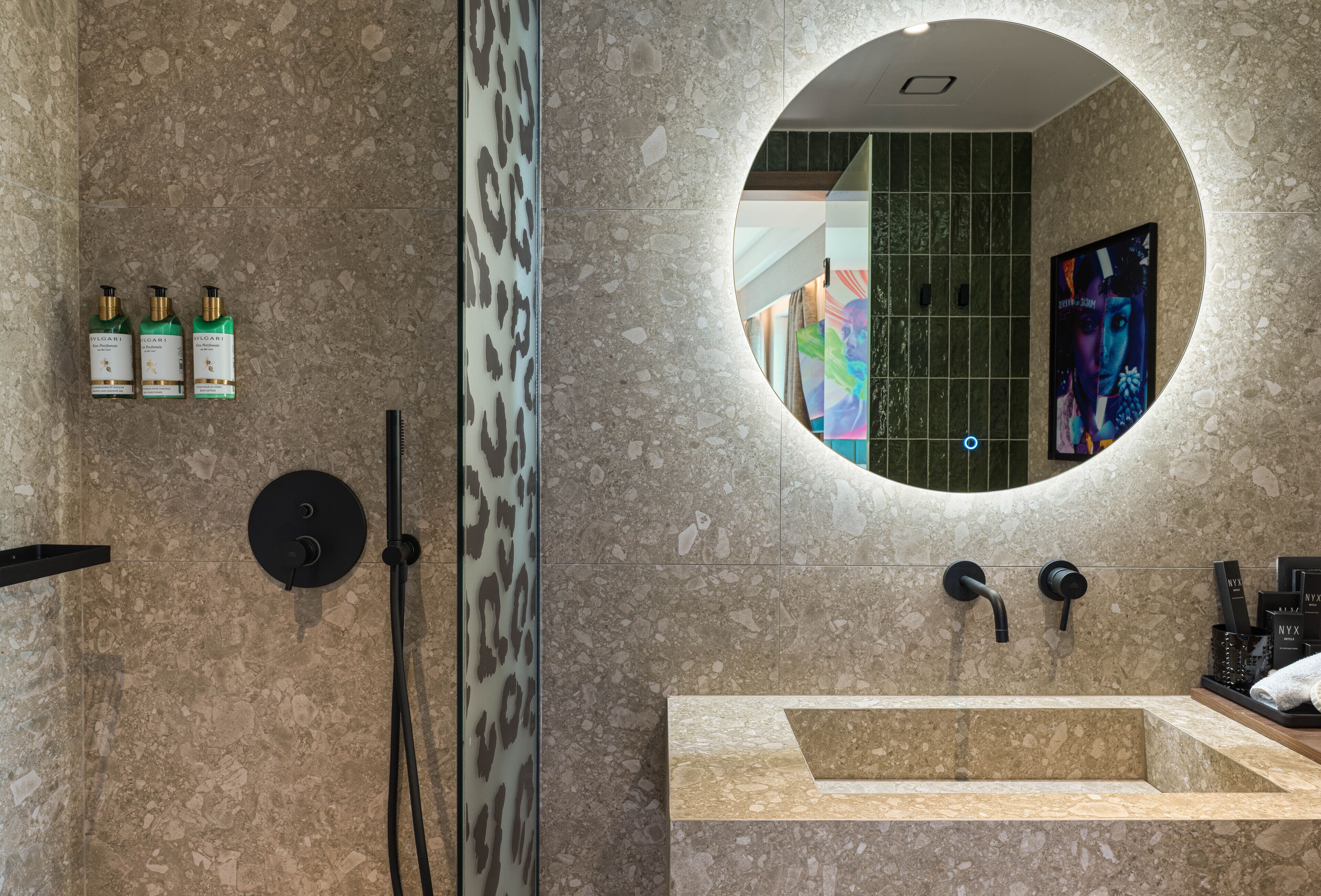 A bathroom with a round mirror, a sink, and three bottles of Bvlgari perfume.