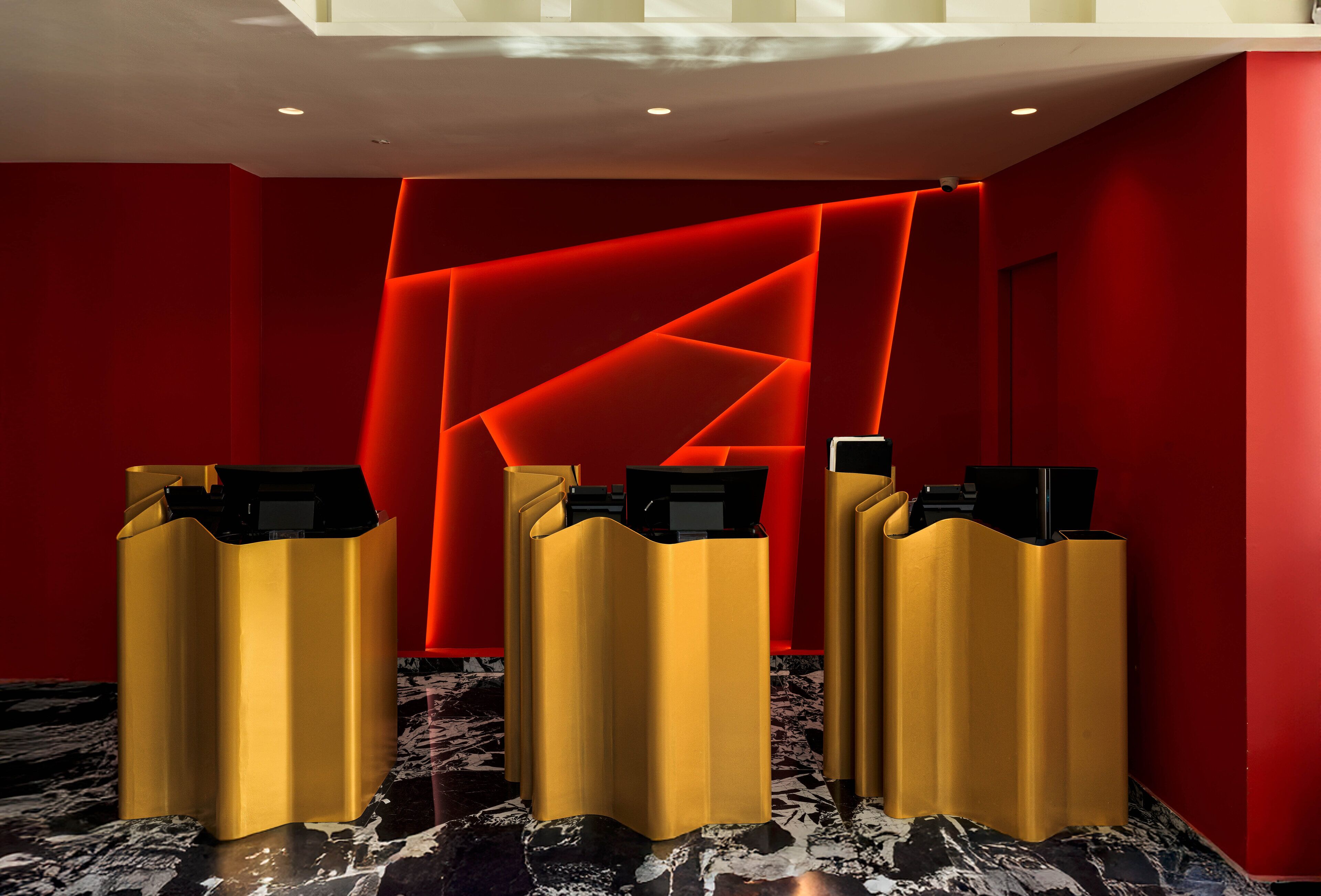 Una zona de recepción con tres mostradores dorados y un diseño mural geométrico rojo.