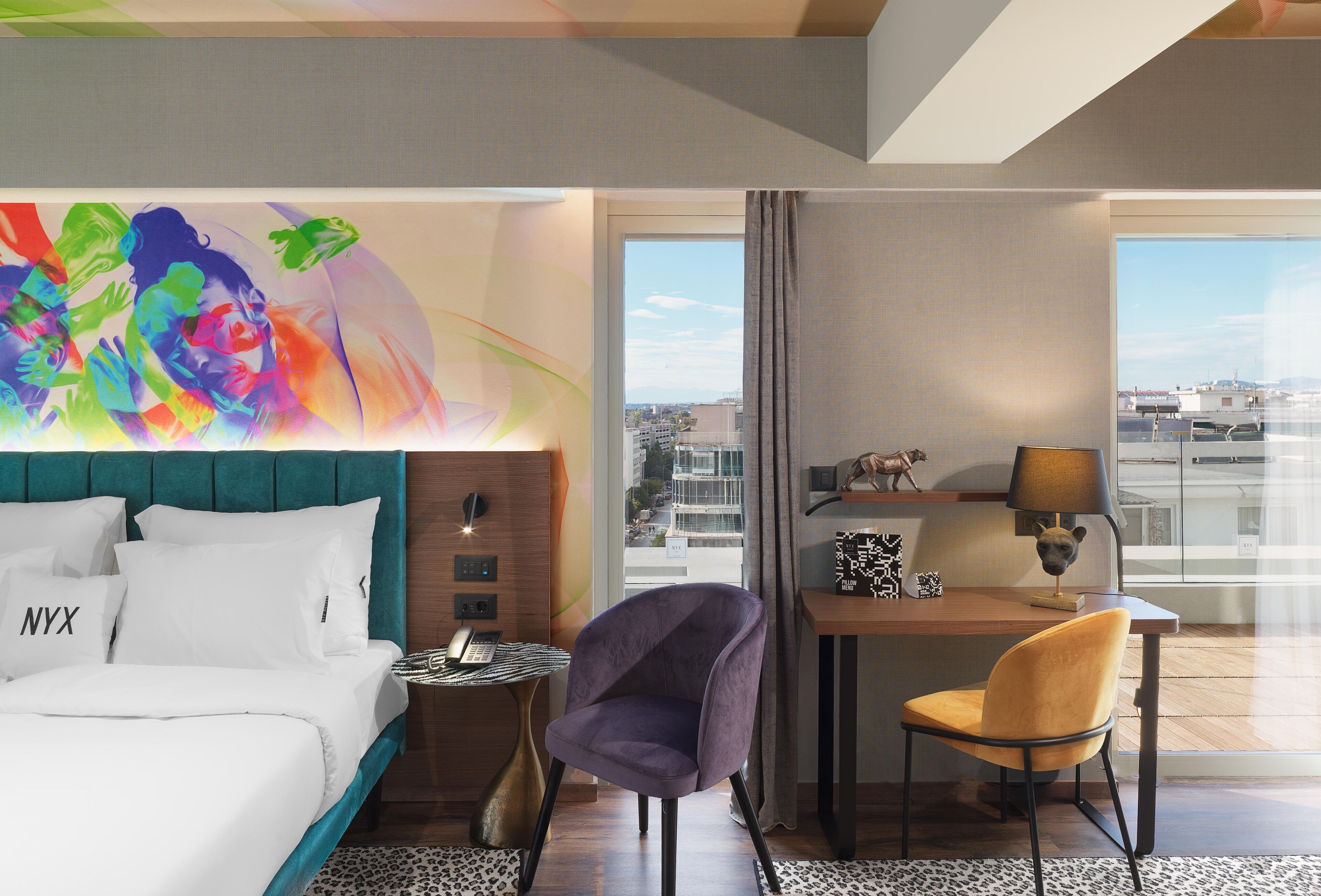 Une chambre d’hôtel avec un lit, un bureau, une chaise, une vue sur la ville et une peinture abstraite aux couleurs vives accrochée au mur.
