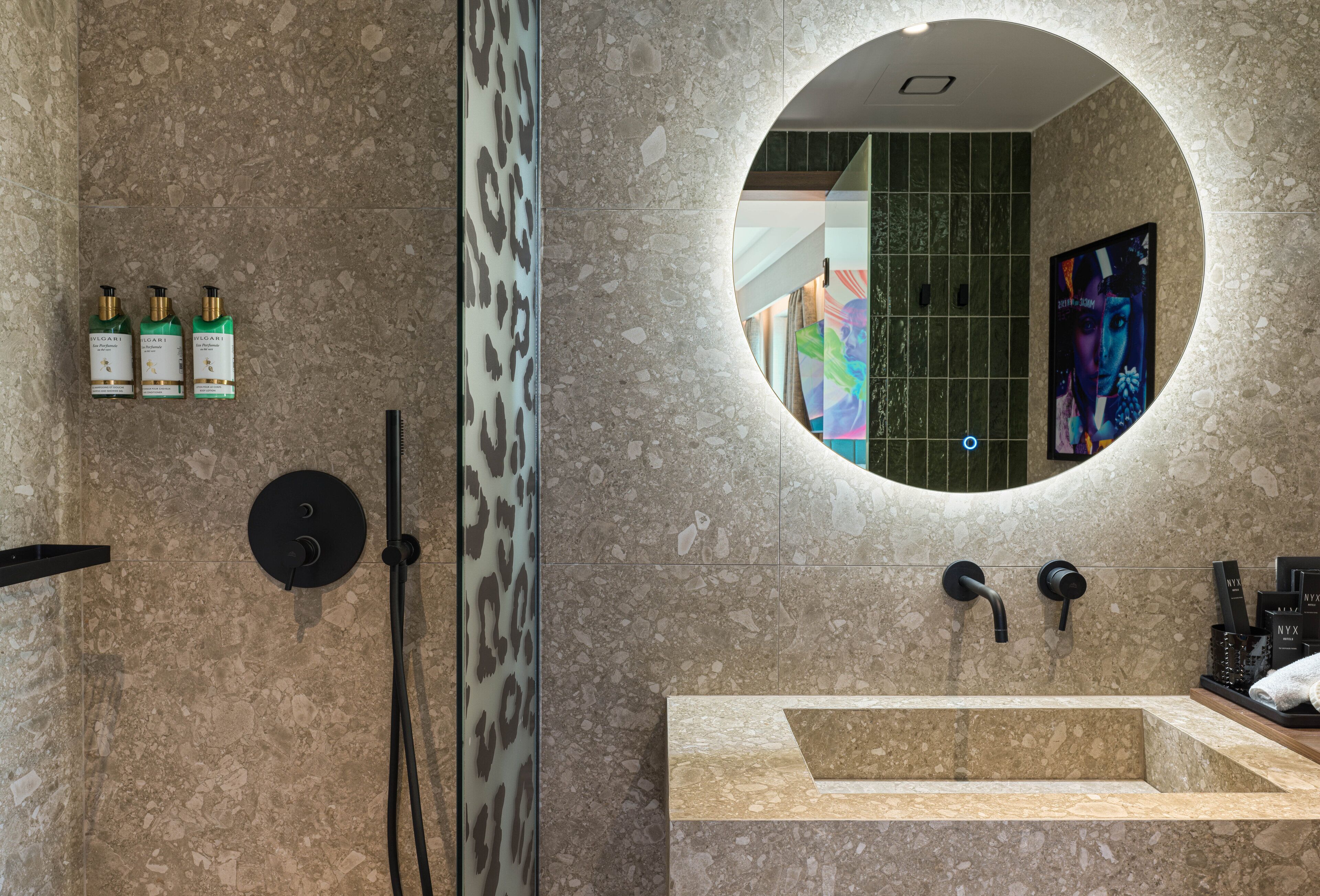 Een moderne badkamer met een ronde spiegel, een wastafel en een douchegedeelte. Drie flessen shampoo staan aan de wand.