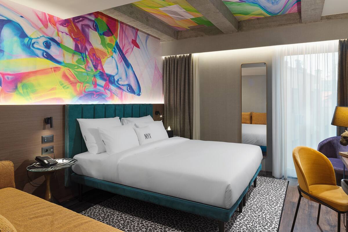 A hotel room with a teal bed, white bedding, a large abstract painting on the wall, a modern lamp, and a leopard print rug.