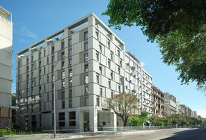 Un hotel de varios pisos con un diseño moderno, que destaca por una entrada imponente con fachada de vidrio y un árbol en primer plano.