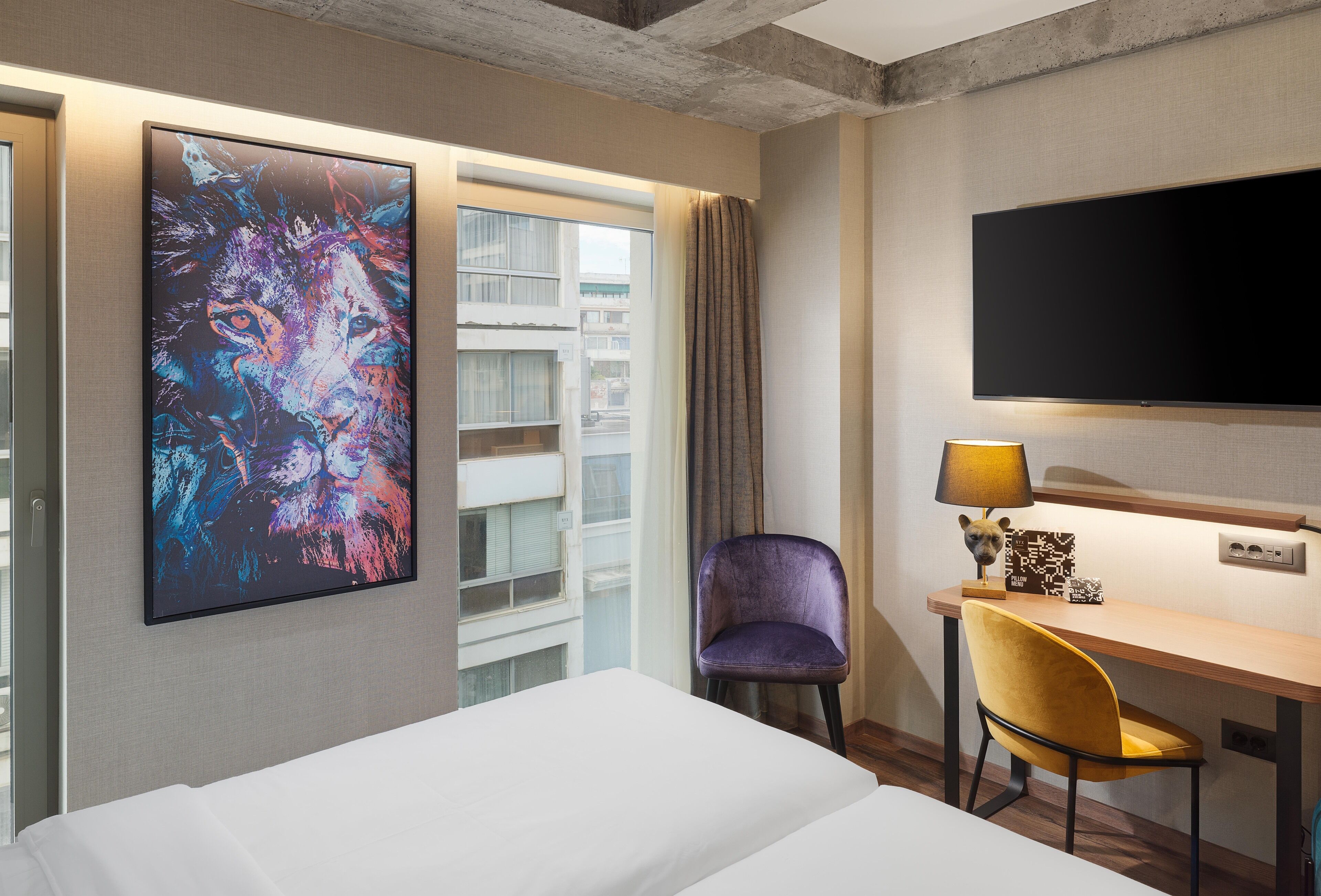 Habitación de hotel con cama, escritorio con lámpara, silla, televisor y un cuadro abstracto grande en la pared.